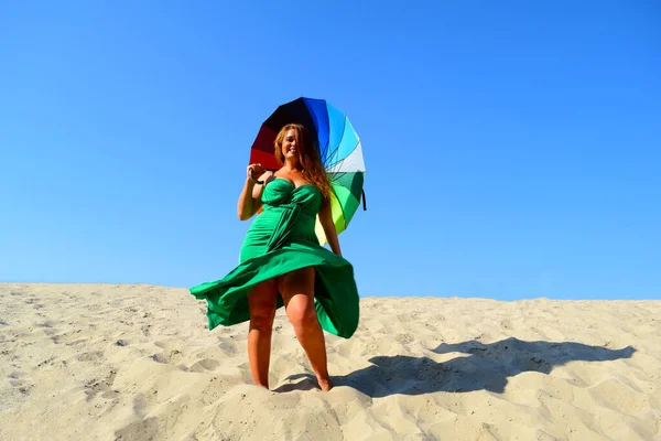Young fat woman in green dress and umbrella stands on the sand
