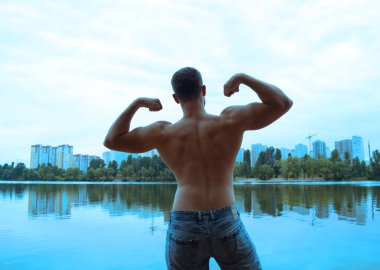 Göl kenarında kollarını kaldırmış ve şehrin arka planındaki evlere yaslanmış genç bir adamın arka görüntüsü.