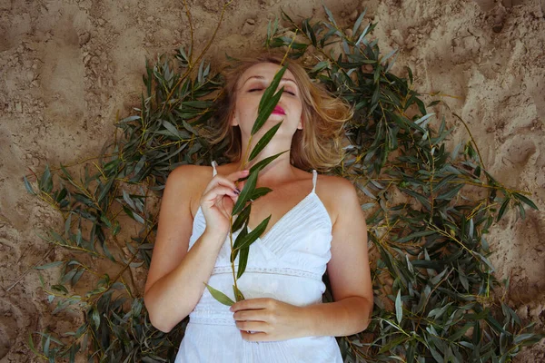 Top view of a woman in a white dress with a long branch with green leaves on her face lies on the sand