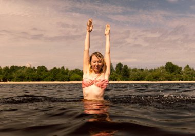 Genç sarışın kadın nehirde suyun içinde dururken ellerini kaldırır. Mayolu kız.