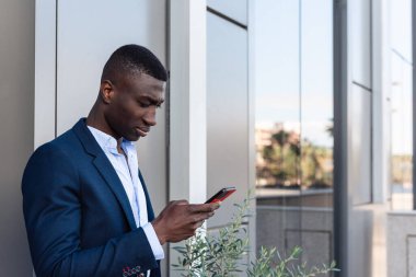 Genç Afrikalı Amerikalı iş adamı modern ofis binasının yakınında akıllı telefon kullanıyor.