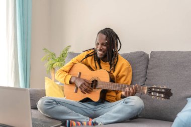 Gülümseyen Afrikalı Amerikalı adam kanepeye oturmuş dizüstü bilgisayarda gitar çalışıyor.