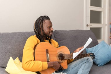 Afro-Amerikalı bir adam yeni bir şarkı öğreniyor gitar akorları çalışıyor kanepede oturmuş nota okuyor.
