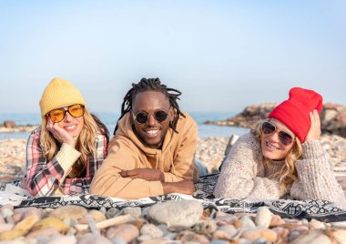 Kumsalda battaniyeye uzanmış kameraya gülümseyen mutlu çok ırklı arkadaşlar.