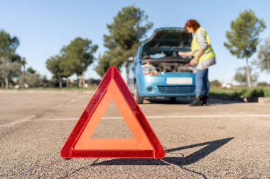 Uyarı üçgeni yola yerleştirildi. Kadın güvenlik yeleği giyiyor. Araba motorlarını kontrol ediyor.