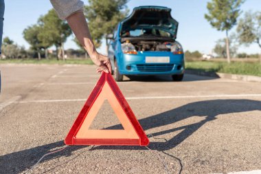 Kadın el, park yerinde kapüşonlu bozuk arabanın arkasına uyarı üçgeni yerleştiriyor.
