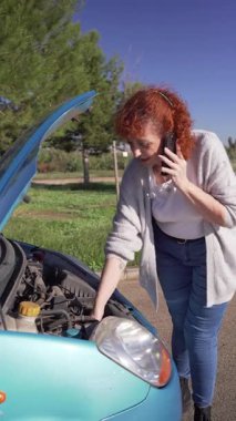 Stresli kızıl saçlı kadın, bozuk kapüşonlu arabanın yanında yol yardımı istiyor.