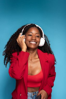 Kırmızı ceketli mutlu İspanyol kadın kulaklıkla müzik dinliyor ve mavi arka planda dans ediyor.