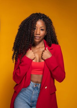 Kırmızı ceketli şık Afro-Amerikan kadın ve sarı stüdyo arka planında poz veren kot.