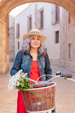 Hasır Şapkalı, kırmızı elbiseli ve Eski Şehir Caddesinde Çiçeklerle Dolu Bisikletli Yaşlı Kadın