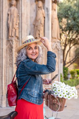 Bisikletli mutlu yaşlı kadın hasır şapka tutuyor ve şehirde kameraya gülümsüyor.
