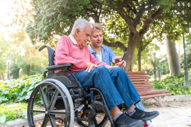 Genç yetişkin torun, tekerlekli sandalyedeki yaşlı adama parkta akıllı bir telefonla yardım ediyor.