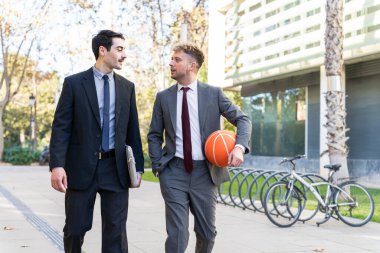 İşten sonra ofisin önünde yürüyen takım elbiseli iki genç işadamı, biri basketbol topu taşıyor.