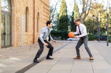 Resmi giyinmiş iki girişimci bir ofis molasında basketbol oynarken eğleniyor.