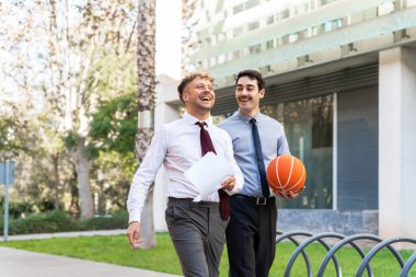 İki iş arkadaşı gülüyor ve dışarıda yürüyor, ellerinde basketbol topu ve iş belgeleri var.