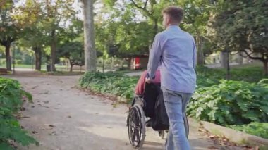 Genç bir adamın yeşil bir parktan tekerlekli sandalyedeki yaşlı bir adamı itmesi.
