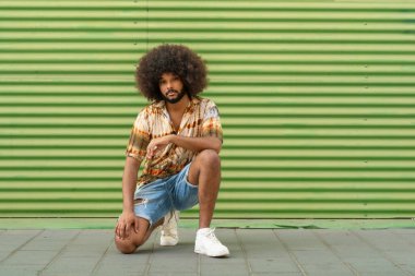 Afro saçlı genç adam şehir ortamında yeşil çizgili duvara yaslanıyor.