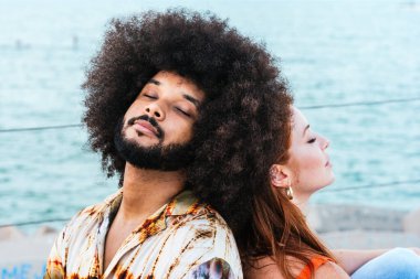 Deniz kenarında sırt sırta oturan ırklar arası bir çift, gözleri kapalı.