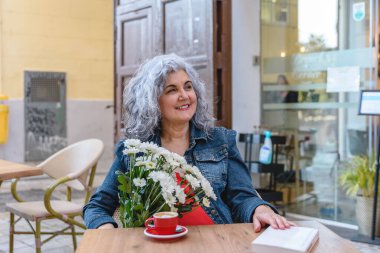 Gri saçlı ve çiçekli gülümseyen son sınıf öğrencisi. Açık kafede, masada kitapla kahvenin tadını çıkarıyor.