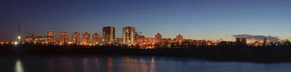 Şehrin kıyı bölgesindeki modern yerleşim yerinin geniş panoramik manzarası. Pencerelerinde ışık ve açık gökyüzü altında parlak sokak ışıkları olan yüksek katlı beton binalar.