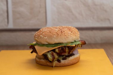 Eritilmiş peynir dilimli dumanlı tavuk burger, domuz pastırması, barbekü sosu, marul, domates ve sarı masa manzaralı susamlı çörek.