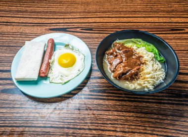 Lezzetli kahvaltı, sahanda yumurta, sosis, kızarmış ekmek ve ahşap bir masada ramen eriştesi, aç bir yemek için mükemmel.