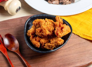 Ahşap bir tahtanın üzerinde kızarmış tavuk parçalarıyla dolu siyah bir kase ve arka planda sarı bir peçete.