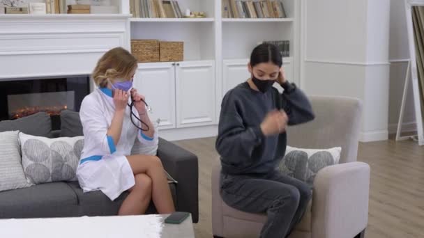 보호가 되는 마스크를 쓰고 있는 젊은 여의사가 가정 간호중에 청진기를 사용하여 환자의 폐를 검사한다 — 비디오