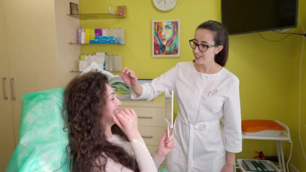 Женщина смотрит в зеркало и обсуждает с косметологом свои проблемы с кожей — стоковое видео