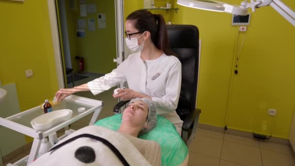 Cosmetologo che applica la crema su faccia di giovane donna durante procedura di cura della pelle in clinica di bellezza — Video Stock