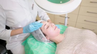 Güzellik uzmanı güzellik kliniğinde kanepede uzanmış genç bir kadının derisini nazikçe temizliyor.