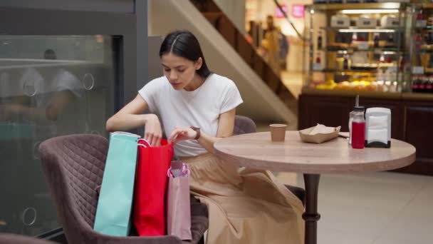 Sitisfied fêmea em roupa elegante verificando suas compras em sacos de papel durante o almoço na praça de alimentação — Vídeo de Stock