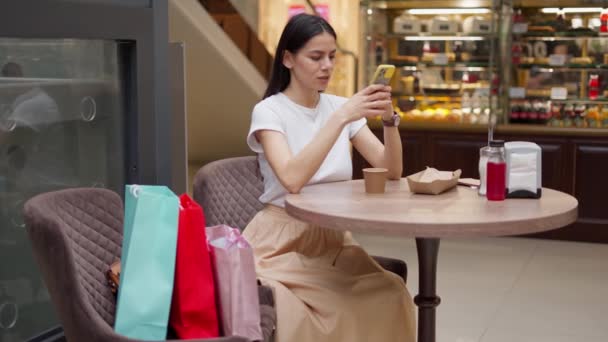 Alışveriş yaptıktan sonra kafede otururken akıllı telefondan arkadaşa fotoğraf gönderen bir kadın. — Stok video