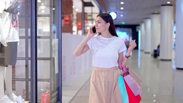 Mujer joven mirando la pantalla de la tienda mientras habla en el teléfono móvil — Vídeos de Stock