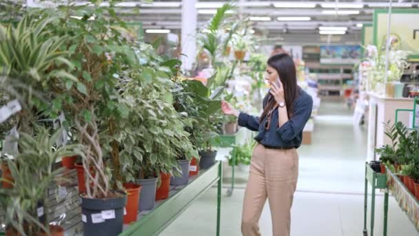 Wanita muda berpakaian santai berbicara di telepon genggam dengan teman saat berbelanja di departemen bunga — Stok Video