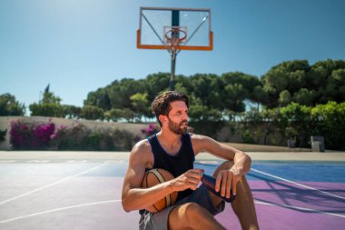 tired basketball player after the match, sits on the basketball field and drinks water clipart
