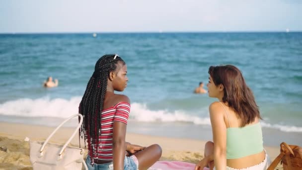 Dwie wielorasowe młode dziewczyny siedzące na piaszczystej plaży — Wideo stockowe