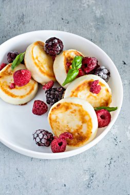 Taze süzme peynir ya da ricotta krepleri (şurup, börek) mavi arka planda taze böğürtlenli beyaz bir tabakta. Lezzetli sağlıklı kahvaltı. Ukrayna mutfağı, Rus mutfağı tatlı yiyecekler.
