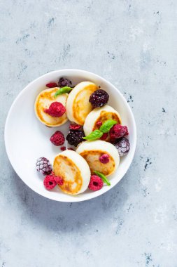 Taze süzme peynir ya da ricotta krepleri (şurup, börek) mavi arka planda taze böğürtlenli beyaz bir tabakta. Lezzetli sağlıklı kahvaltı. Ukrayna mutfağı, Rus mutfağı tatlı yiyecekler.