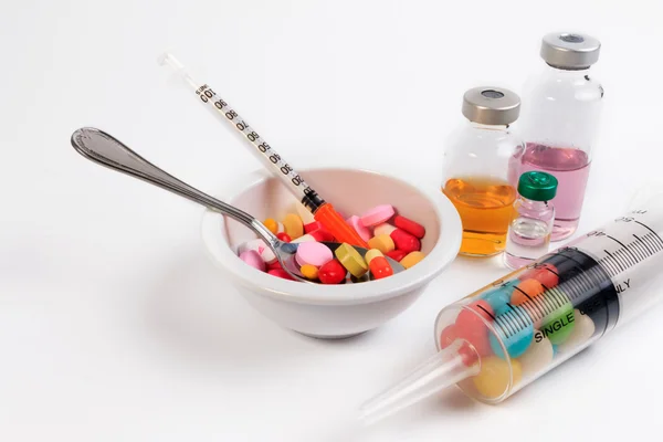 Muchas píldoras de color, cápsulas y jeringa en una taza con una cuchara y un frasco de vacuna . — Foto de Stock