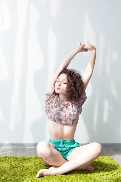 Young girl with curly hair sitting on the floor, on a green rug, do stretching exercises. Doing exercises in the morning in the bright sun room. Pull your hands up. Practice yoga at home. Enjoy life.