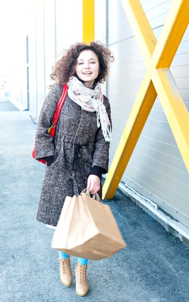Happy girl finishes shopping. In the hands packages with purchases.  Make purchases with pleasure. Girl in a coat in the spring, wavy curly hair is developing the wind, full portrait outdoors growth. — ストック写真