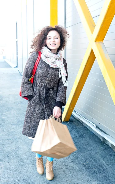 Chica feliz termina de comprar. En las manos los paquetes con las compras. Hacer compras con placer. Chica en un abrigo en la primavera, pelo ondulado rizado está desarrollando el viento, retrato completo crecimiento al aire libre . — Foto de Stock