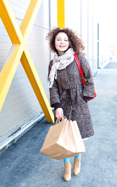 Menina em um casaco na primavera, cabelo encaracolado ondulado está desenvolvendo o vento, retrato completo ao ar livre crescimento. Menina feliz termina de fazer compras. Nas mãos pacotes com compras. Fazer compras com prazer .. — Fotografia de Stock