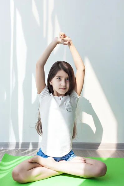Pose de loto. niña haciendo gimnasia en una esterilla verde para el yoga. hacer ejercicio físico y estiramiento en una habitación luminosa. fitness para niños, yoga para niños . — Foto de Stock