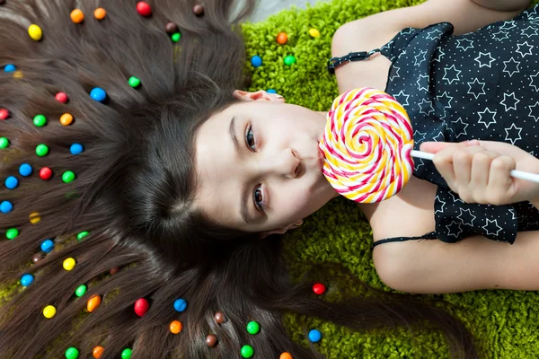 Little girl with dark hair lying on the floor among the sweets. Top view of a big yellow and pink lollipops in my mouth. Many candies in her hair. fun and excitement on the face of the child.
