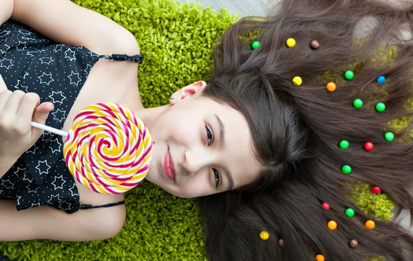 Kleines Mädchen mit dunklen Haaren, das auf dem Boden zwischen den Süßigkeiten liegt. Draufsicht auf einen großen gelben und rosa Lutscher in den Händen. Lächeln, Freude und Emotion auf dem Gesicht des Kindes. viele Bonbons im Haar. — Stockfoto