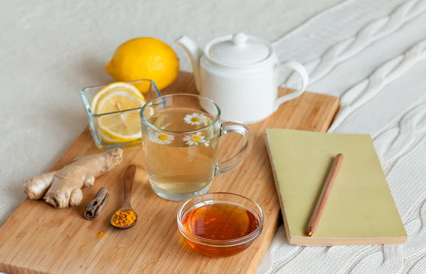 Té de hierbas en taza de vidrio con flores de manzanilla, cúrcuma y miel en una tabla de madera. Tratamiento con una bebida fría. Tratamiento de remedios caseros en la cama. Lectura de libros — Foto de Stock