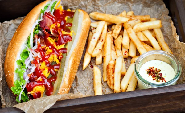 Grote smakelijke hotdog met saus en frietjes op het perkament op de houten achtergrond. hotdogs op gourmet. — Stockfoto