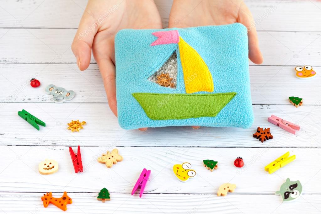 Childrens toy made of colored fleece for the development motor skills in the hands women. Pouch with a ship the filled plastic beads and figurines on white wooden background. handmade toys.